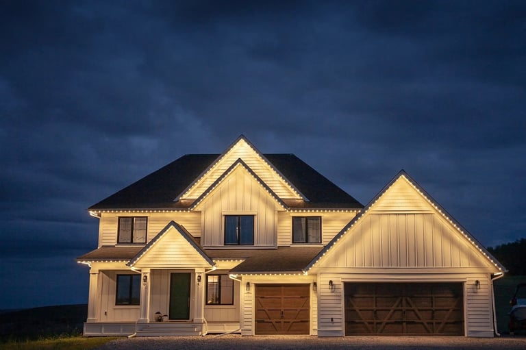Illuminated two-story cream-colored house with peaked gable roofs, dark trim, and attached garage at dusk against a stormy blue sky
